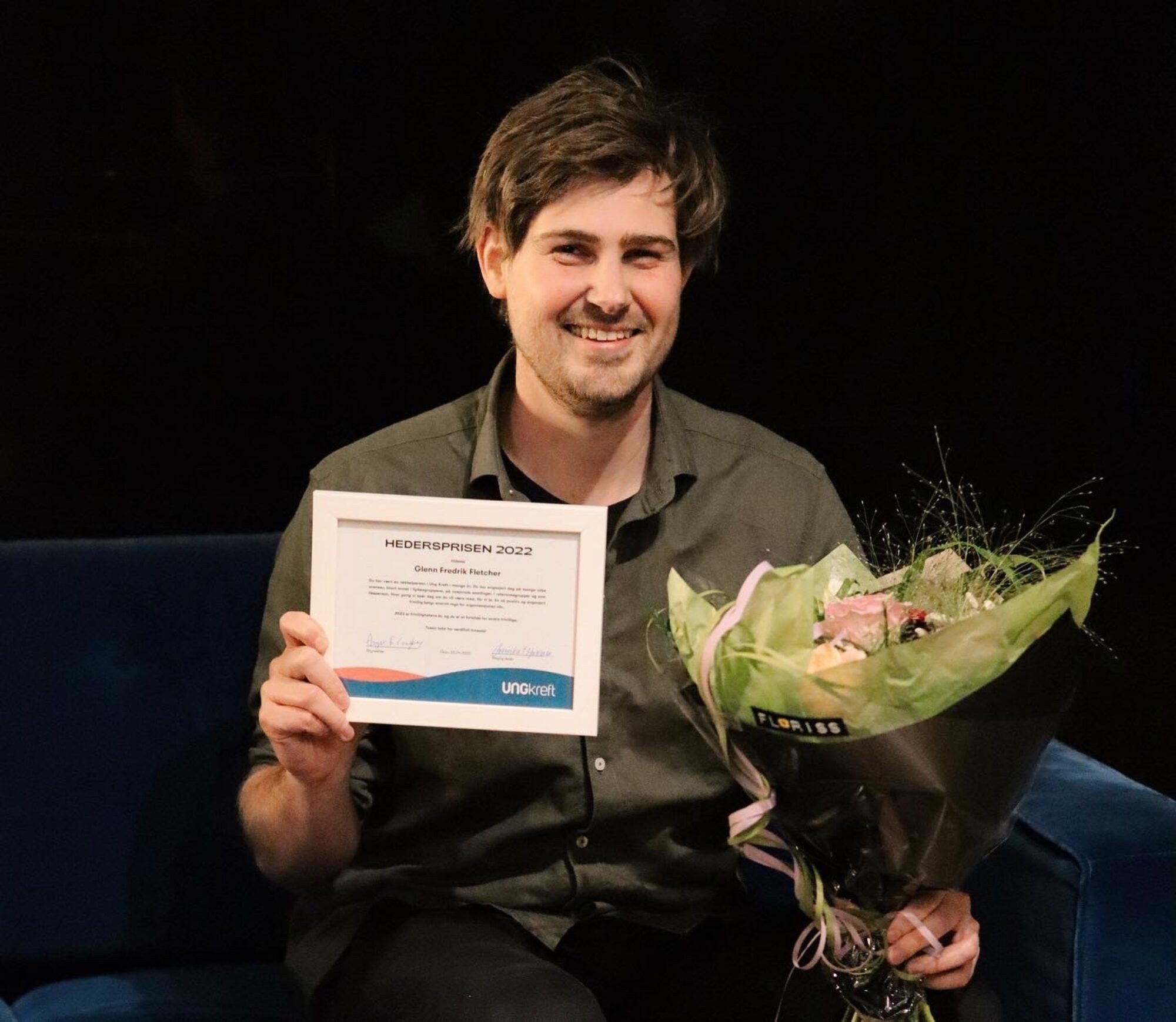 Hedersprisvinner Glenn sitter og smiler til kamera med diplom og blomster i hånden.